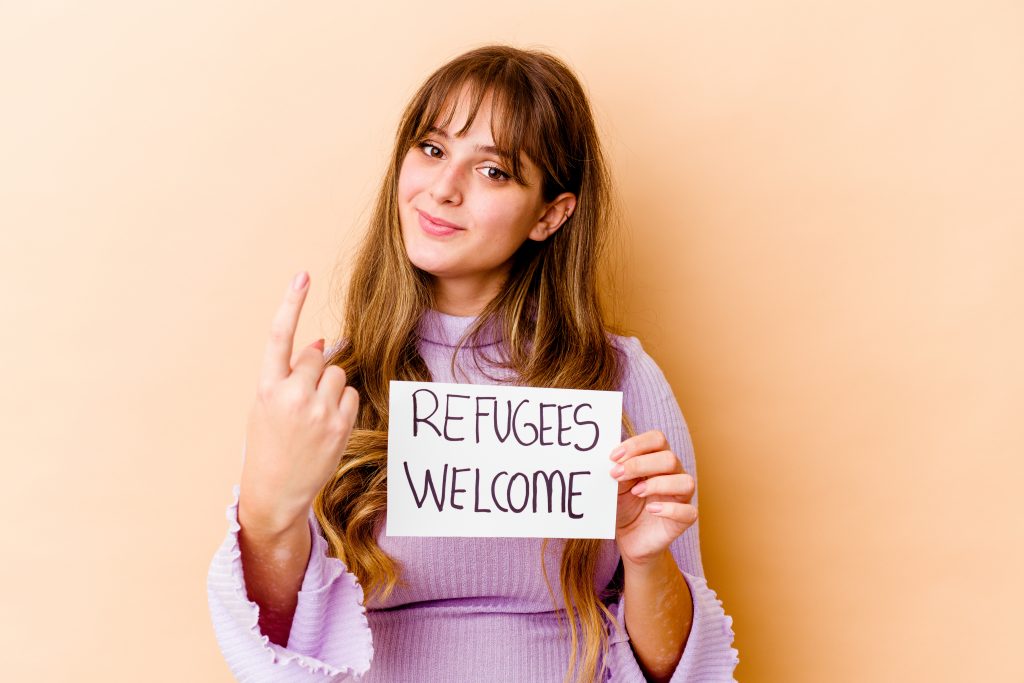 young caucasian woman holding refugees welcome placard isolated pointing with finger you as if inviting come closer