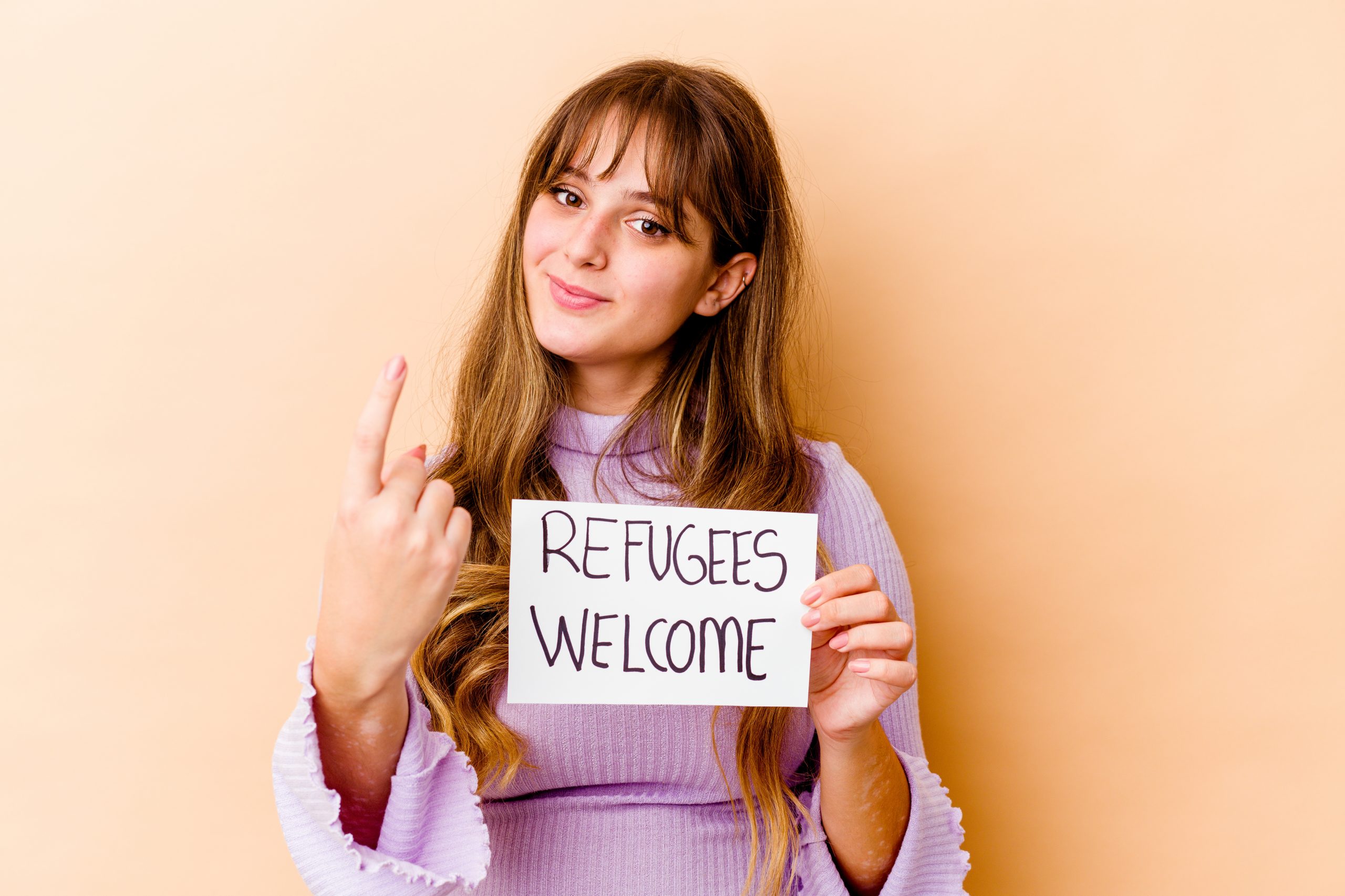 young caucasian woman holding refugees welcome placard isolated pointing with finger you as if inviting come closer scaled